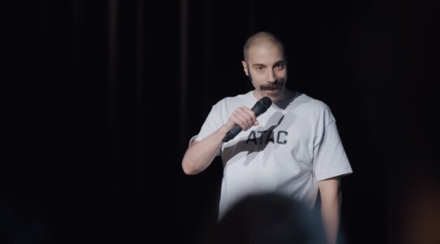 A white man with a large mustache performs stand-up comedy under a spotlight.